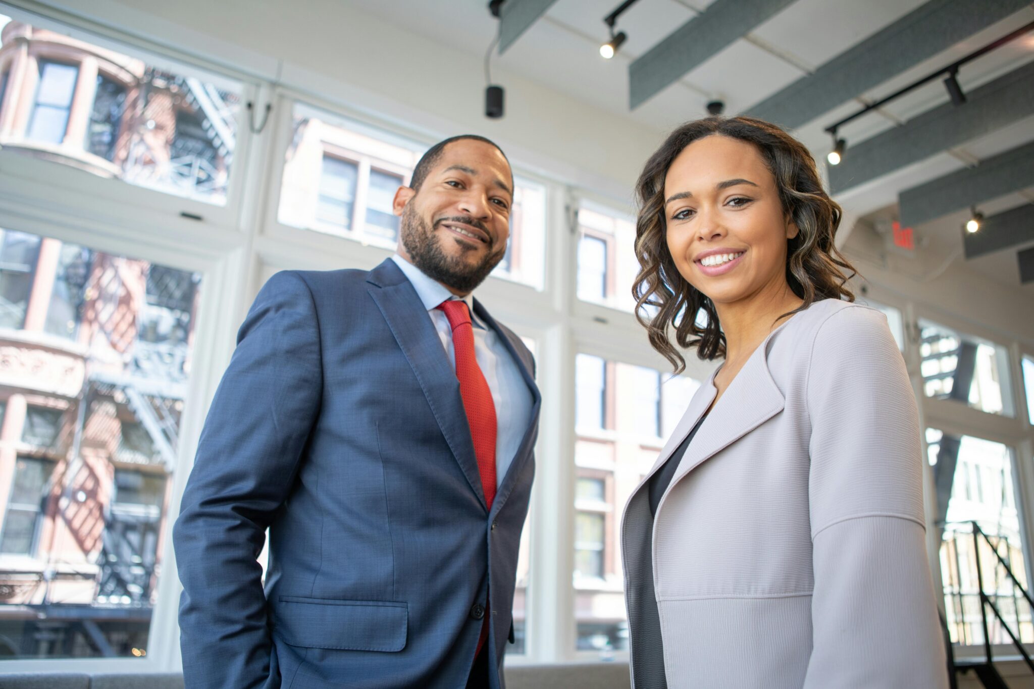 Two professionals in business attire smiling confidently inside a modern office with large windows.