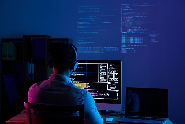 A person wearing headphones sits at a desk with multiple computer screens displaying code