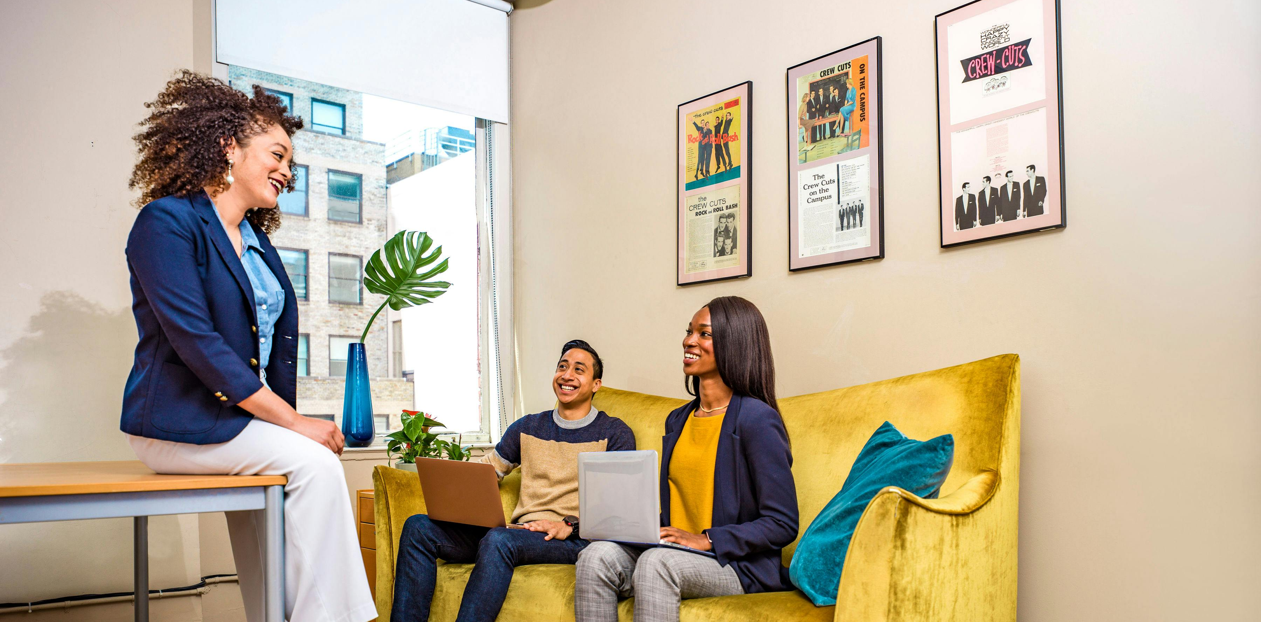 Office team meeting with three people, two sitting on a yellow couch with laptops and one woman sitting on a bench near a window with a blue vase and green leaf